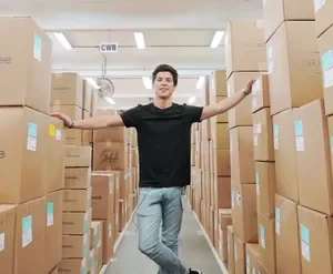 Homem com expressão de sucesso em frente a uma coleção de roupas de marca, vestindo um casaco elegante, com fundo de estúdio moderno e luz suave.